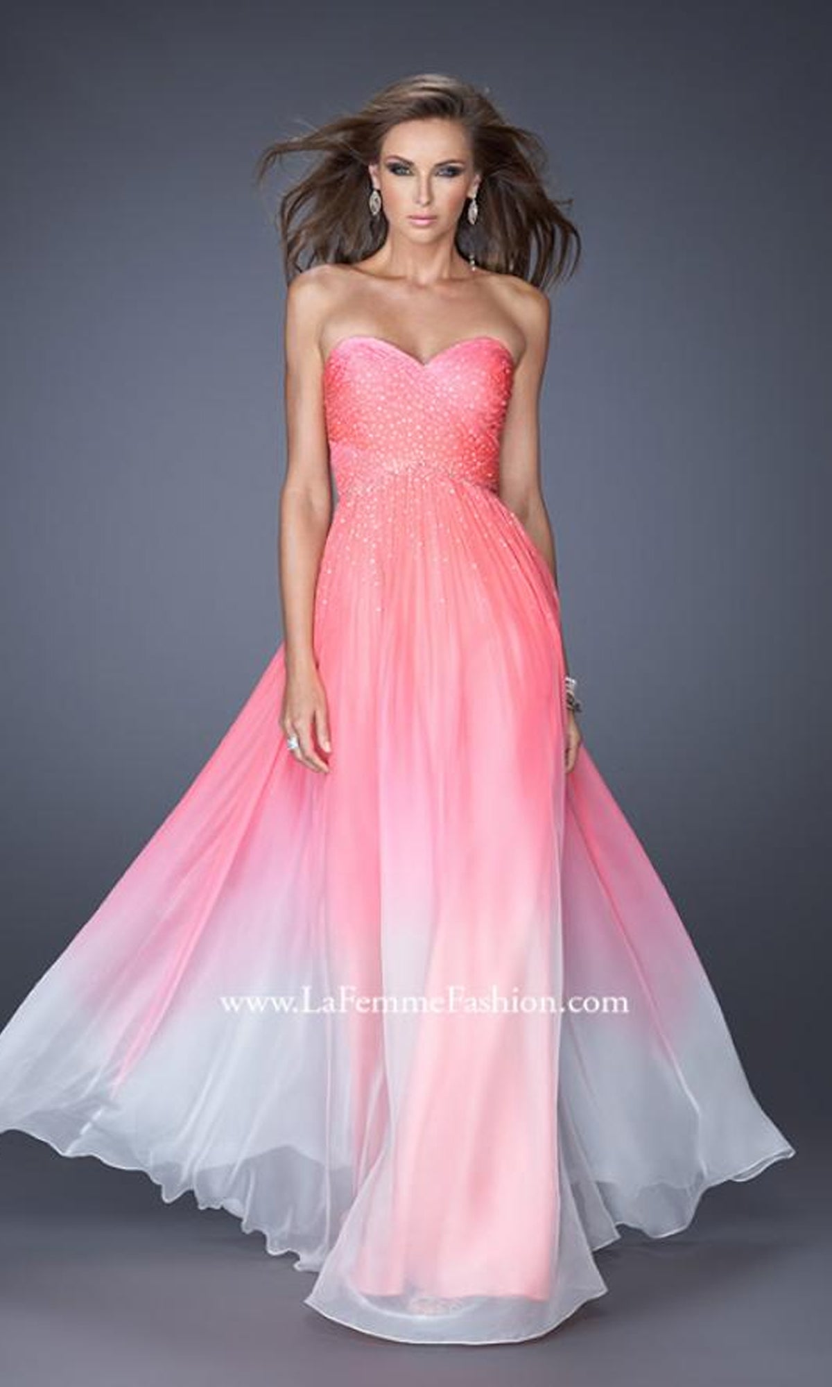 A woman stands against a plain gray background wearing the Floor Length La Femme Formal Dress—a strapless, sweetheart neckline gown with a pink-to-white ombre, fitted beaded bodice and flowing skirt.